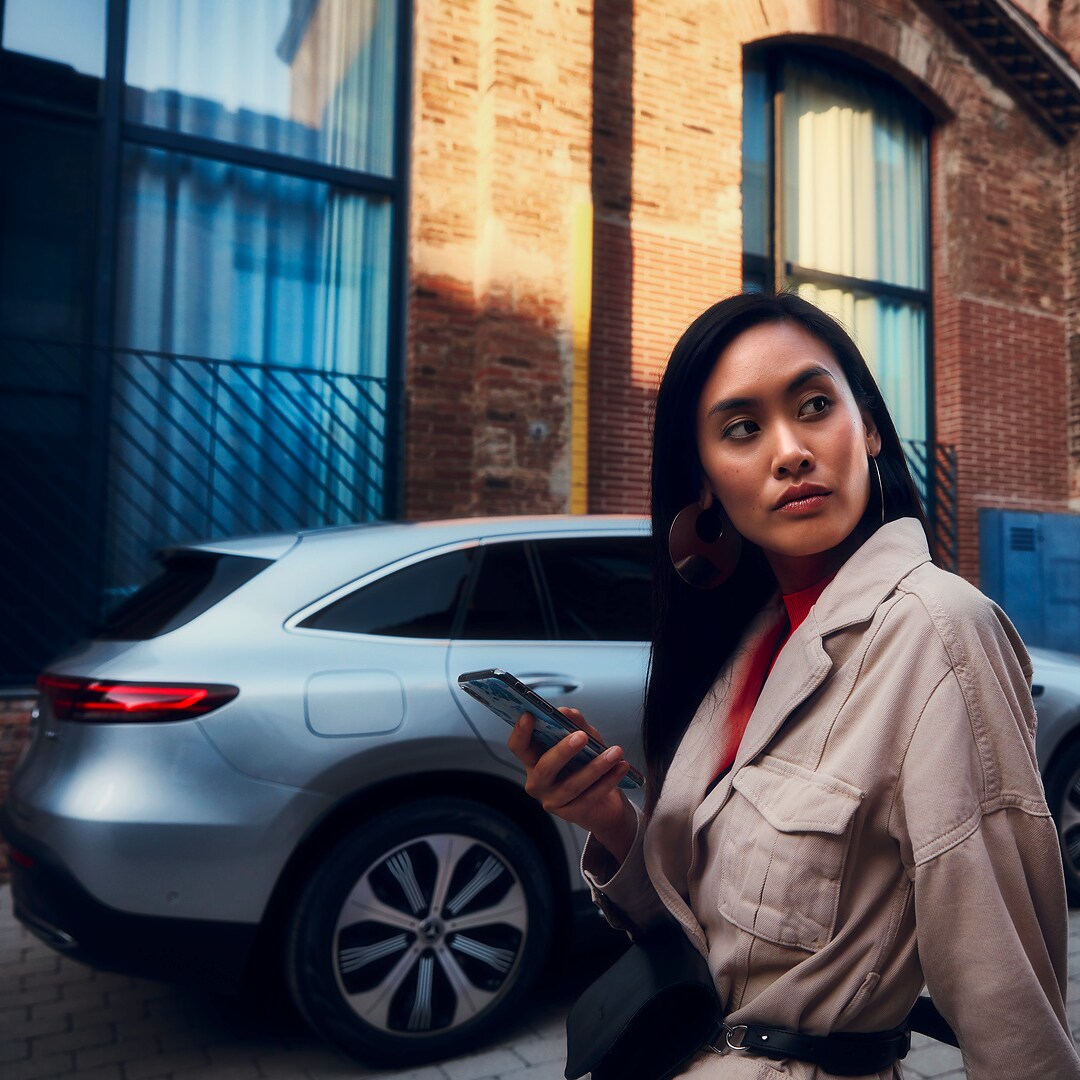 A woman standing in front of her Mercedes with a smartphone in her right hand.