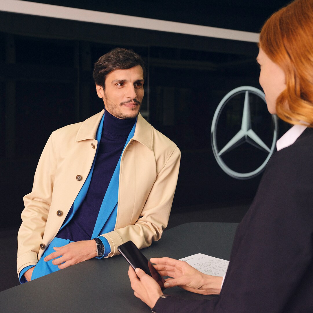 A customer leaning at the counter turns expectantly to face a Mercedes-Benz employee.